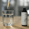 A glass of clear water on a wooden surface stands beside a dark bottle labeled KÄÄPÄ Mushrooms Reishi Extract, as a dropper dispenses this natural sleep aid from KÄÄPÄ Mushrooms into the water.