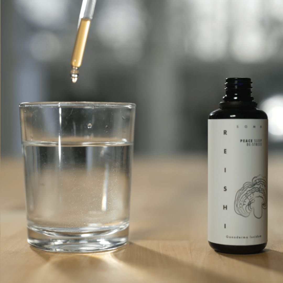 A glass of clear water on a wooden surface stands beside a dark bottle labeled KÄÄPÄ Mushrooms Reishi Extract, as a dropper dispenses this natural sleep aid from KÄÄPÄ Mushrooms into the water.