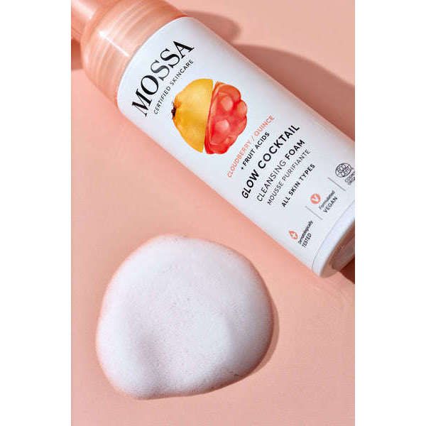 A bottle of Mossa Glow Cocktail Cleansing Foam by Mossa rests on a pink surface next to a dollop of white foam. The vitamin-rich formula with fruit images is labeled for all skin types and promises glowing, radiant skin.