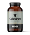 A brown glass jar labeled DENSE Colostrum, a 135g dietary supplement from DENSE, features a black cow icon and "Colostrum Powder from Raw Milk" text on a light background.