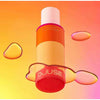 A vibrant orange and yellow bottle of Djusie Liquid Silk Perfect Cleansing Oil by Djusie appears to float above a peach-pink background, surrounded by glistening droplets—ideal for removing water-resistant makeup.
