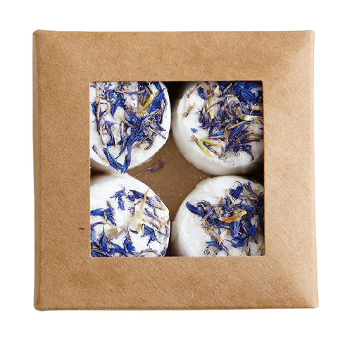 A brown cardboard box with a square window reveals four round, white Kaurilan Sauna Eucalyptus Footbath bombs by Kaurilan Sauna—topped with dried blue and yellow flower petals, handmade in Helsinki from vegan, natural ingredients.