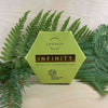 A hexagonal green box of Luonkos Infinity Oil Cleansing Cake stands upright on a light surface, surrounded by green fern and pine branches—ideal for sensitive skin.