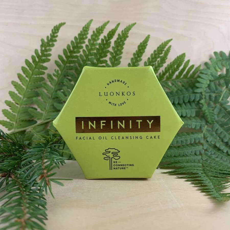 A hexagonal green box of Luonkos Infinity Oil Cleansing Cake stands upright on a light surface, surrounded by green fern and pine branches—ideal for sensitive skin.