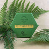 A green hexagonal box labeled Luonkos Vitality Body Oil Cake by Luonkos rests on light wood, surrounded by fern leaves. The packaging features Forest Microbe Extract and the Reconnecting Nature logo.