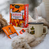 Wrapped in warm winter clothes, someone pours a hot drink from a thermos into a Snufkin mug beside a box and packets of Nordqvist All Things Fun Are Good For Your Tummy rooibos tea, all set on snow.
