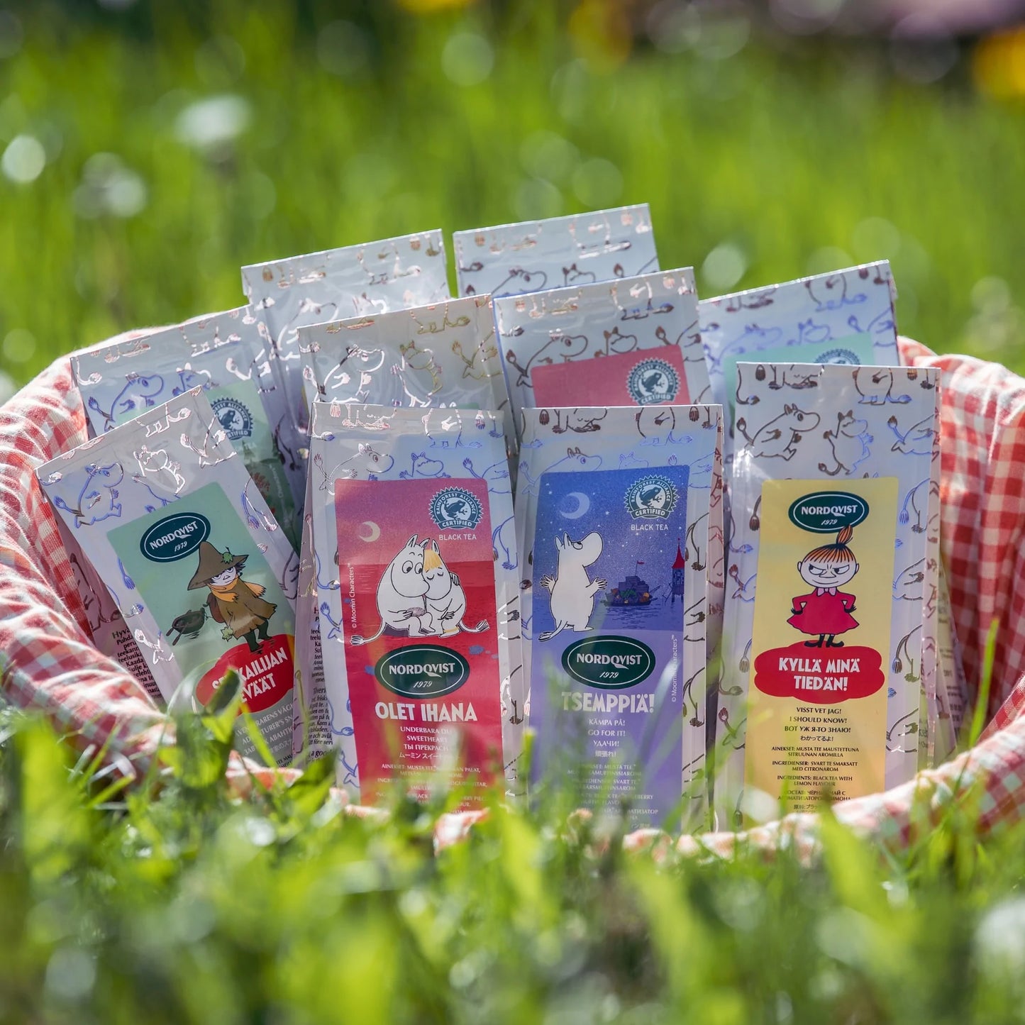 A basket lined with red and white checkered cloth holds colorful Nordqvist Moomin Go for It! tea packages, featuring Moomin characters, set outdoors on bright green grass in sunlight.