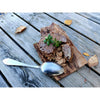 A rustic Finnish treat, Pielisen Kala Finnish Fish Pasty by Pielisen Kala is served on bark with a silver spoon and autumn leaves, capturing the authentic charm of traditional Muikkukukko flatbread and fish.