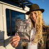 A person with long blond hair and a hat smiles while holding a Renjer Nordic Elk Jerky Black Pepper packet in front of a vehicle. The label shows the brand Renjer and highlights its 100% natural, traditionally Nordic qualities.