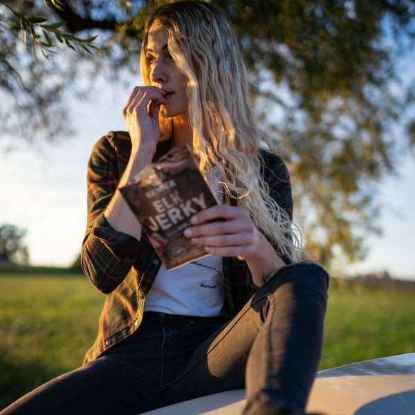 A woman with long blonde hair enjoys Renjer Nordic Elk Jerky Black Pepper outdoors on a sunny day. Wearing a plaid shirt and jeans, she savors the flavorful snack surrounded by trees and grass.