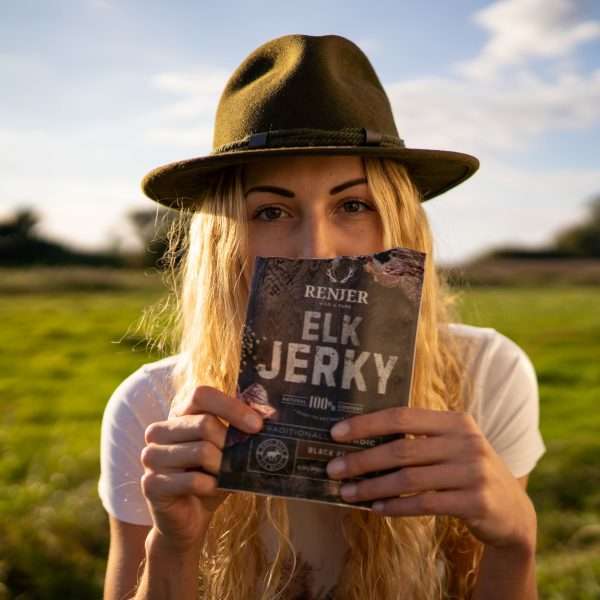 A person with long blonde hair in a green hat and white shirt holds Renjer Nordic Elk Jerky Black Pepper by Renjer in front of their face, standing outdoors in a grassy field beneath a blue sky.