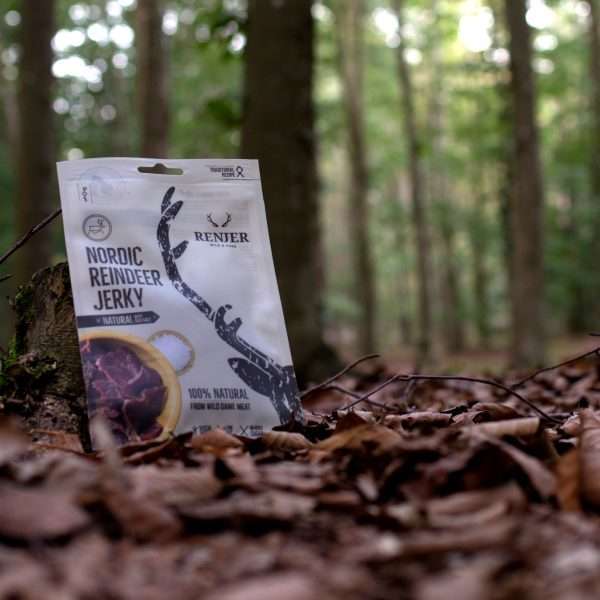 A packet of Renjer Nordic Reindeer Jerky Sea Salt rests against a tree stump on a leaf-strewn forest floor, with blurred green trees in the background.