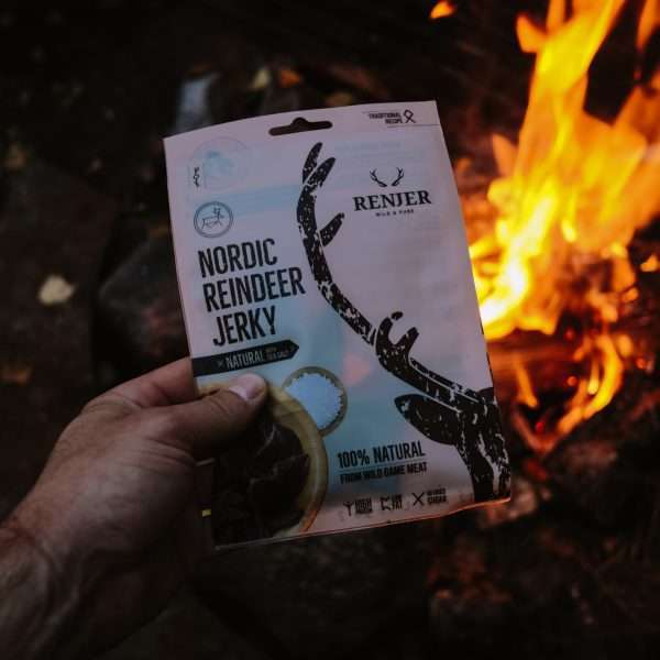 A hand holds a pack of Renjer Nordic Reindeer Jerky Sea Salt by Renjer near a campfire. The packaging shows antler graphics and highlights 100% natural Lapland reindeer meat and wild game expertise.