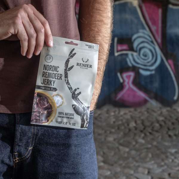 Someone holds a pack of Renjer Nordic Reindeer Jerky Sea Salt from Renjer in front of a graffiti-covered wall on rocky ground, highlighting the distinct flavor of Lapland reindeer meat in an urban environment.