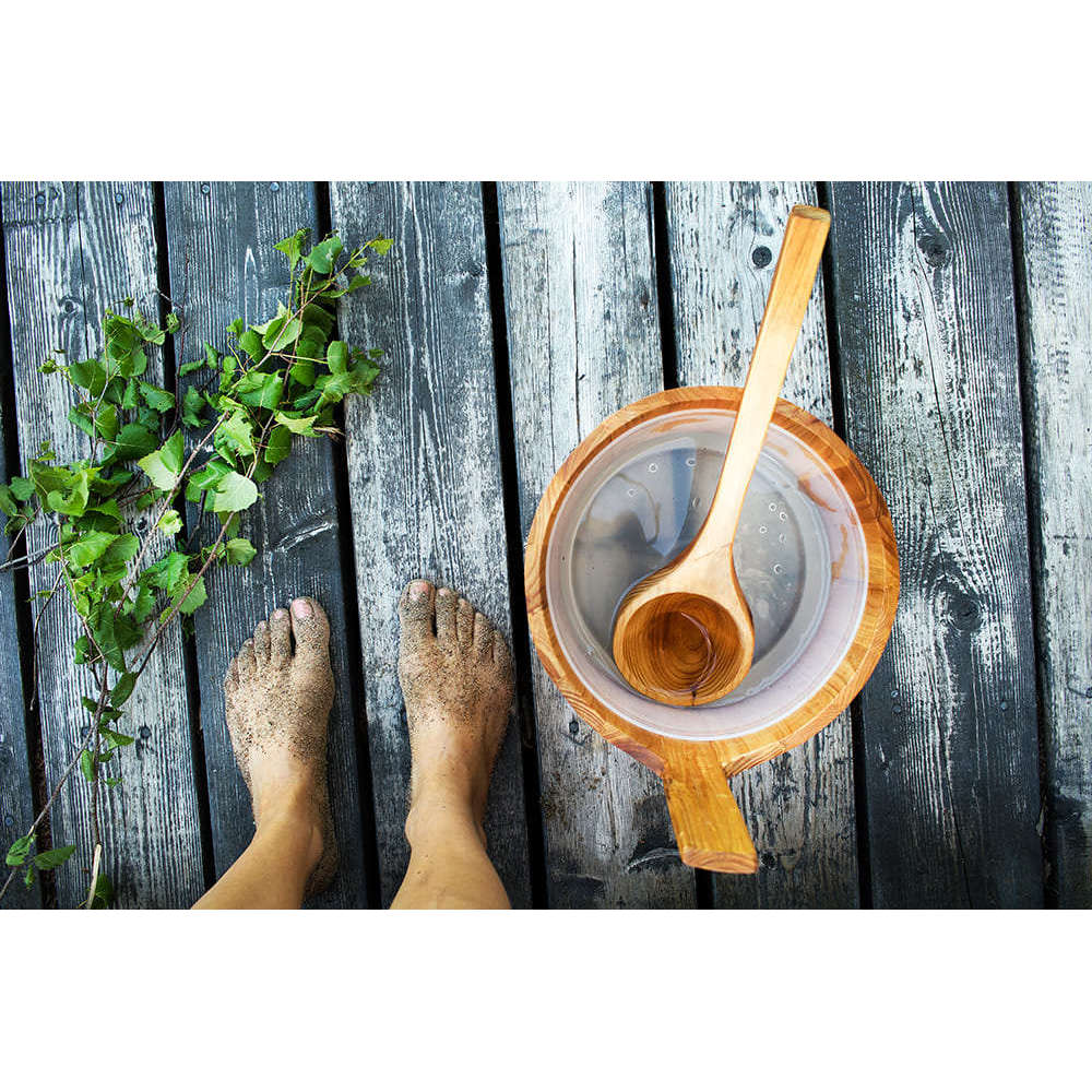 Sandy bare feet on weathered planks rest beside a wooden bucket and ladle, with an Emendo Sauna Fragrance (Eucalyptus, Birch, Smoke Sauna & Tar) set to fill the air with refreshing scents and essential oils.