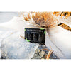 An Emendo Sauna Fragrance Salmiac, Pine & Sisu box by Emendo, made in Finland, rests outdoors on cracked ice among dried reeds with sunlight shining behind.