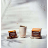 A cup of coffee is placed between a packet and a box of Four Sigmatic Instant Mushroom Coffee Lions Mane by Four Sigmatic, both set on stone blocks against a light textured background.