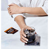 A person in a white shirt sits at a table, holding a glass of iced Four Sigmatic Instant Mushroom Coffee Lions Mane. On the table is a Four Sigmatic packet labeled Organic and a single blueberry on a coaster.