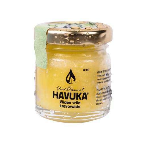 A small, gold-lidded glass jar of Havuka Five Herbs Facial Balm by Havuka. This yellow Finnish face and eye balm features a green seal and water droplets and is enriched with wild herbs from Finland.