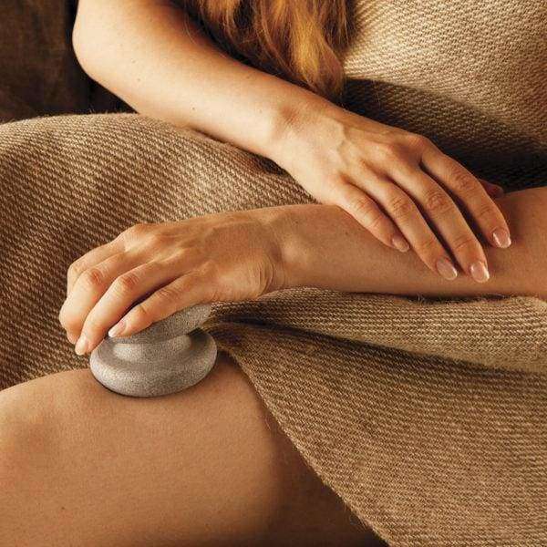 A person draped in a brown textured cloth uses the Hukka Sauna-Joy Massage Aid by Hukka on their thigh, while their other hand rests gently on their lap.