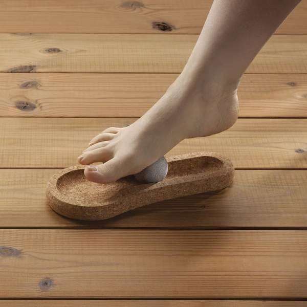 A person rolls the Hukka Solejoy, a gray massage stone by Hukka, on a foot-shaped cork base on a wooden floor—ideal for foot reflexology.
