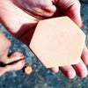 A hand holds the Luonkos Delicious Body Cake—a peach-colored, hexagonal soap bar by Luonkos—while bare feet and another bar rest nearby on a rough outdoor surface.