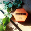A hexagonal orange box labeled Luonkos Delicious Body Cake rests on wood beside green leaves in sunlight, evoking the nourishing care and essential oils of this natural body moisturizer by Luonkos.