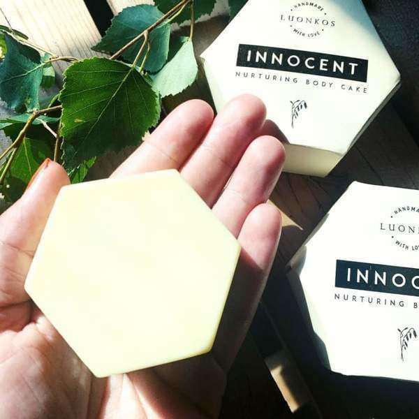 A hand holds a hexagonal, pale yellow Luonkos Innocent Nurturing Body Cake for dry and sensitive skin. Two packaged bars and green leaves appear in the background.