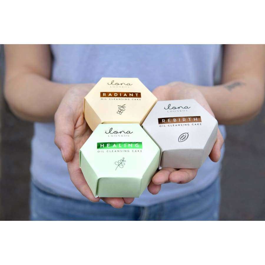 A person holds three hexagonal boxes labeled Radiant, Healing, and Rebirth from Luonkos, each containing the vegan Luonkos Rebirth Oil Cleansing Cake in pastel packaging.
