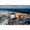 A jar of Biomed Reindeer Bone Broth Powder + Curcumin + Ginger + Chili and a wooden cup sit on a rocky surface outdoors, with hills and sky softly blurred in the background.