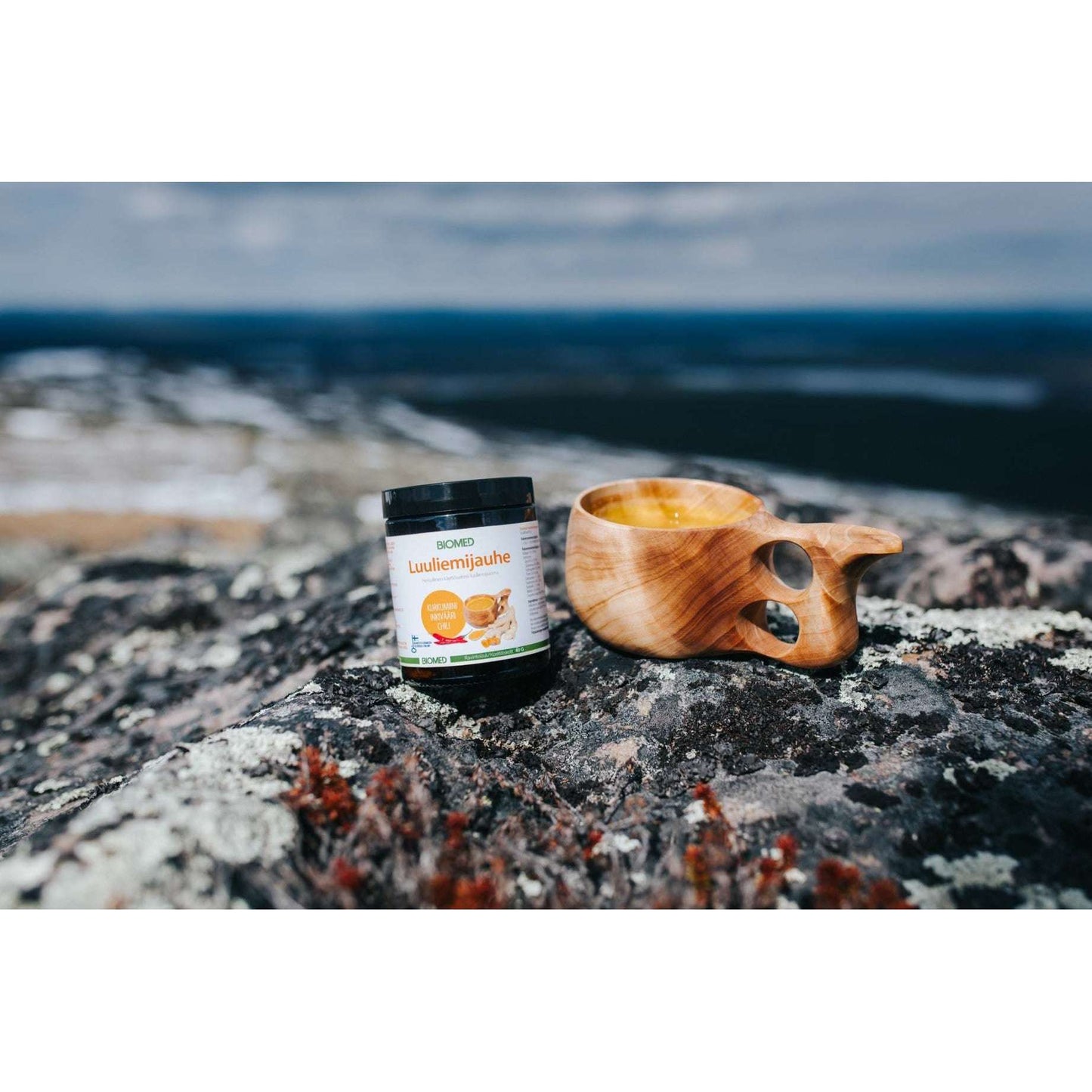 A jar of Biomed Reindeer Bone Broth Powder + Curcumin + Ginger + Chili and a wooden cup sit on a rocky surface outdoors, with hills and sky softly blurred in the background.