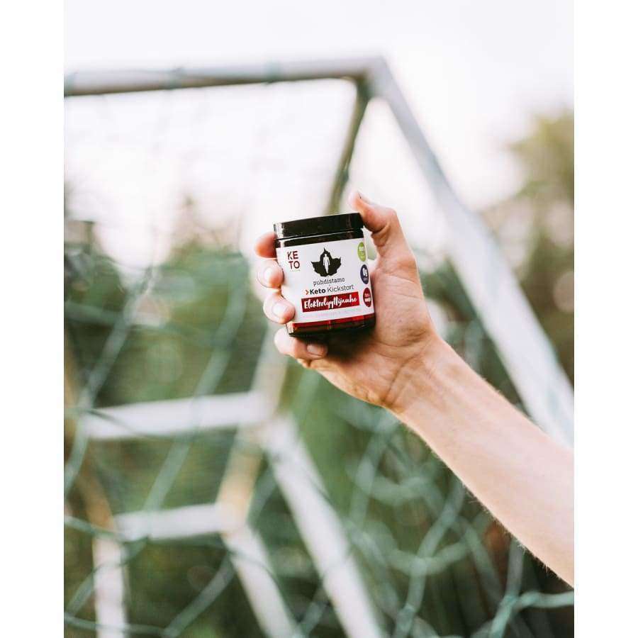 A hand holds a small jar labeled “Puhdistamo Electrolyte Powder Forest Fruit” by Puhdistamo in front of a soccer goal net outdoors, suggesting its support for hydration, with the background softly blurred.