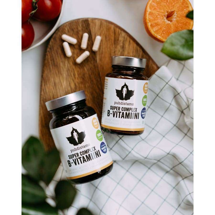 Two bottles of Puhdistamo Super Complex B-complex from Puhdistamo rest on a white surface with a checkered cloth, surrounded by capsules, tomatoes, an orange half, and green leaves.