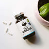 A bottle of Puhdistamo Triple Magnesium by Puhdistamo rests on a white surface beside three white capsules and a potted green plant, showcasing this highly absorbable supplement ideal for supporting heart health.