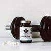 A jar of Puhdistamo Triple Magnesium by Puhdistamo rests on a white surface beside a black dumbbell, with several white capsules scattered close by.