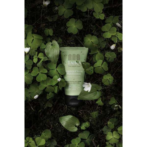 A green tube of Taika Day Cream by Taika rests among clover leaves and small white flowers, emphasizing its vegan skincare formula.